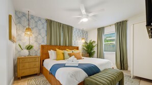 Une chambre à coucher bien rangée, avec un lit, des tables de chevet, un ventilateur de plafond et une fenêtre avec des rideaux.
