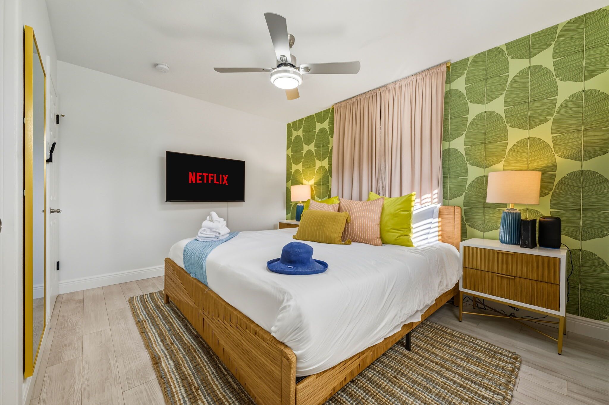 A bedroom with a bed, a ceiling fan, a wall-mounted TV, a bedside table with a lamp, and a patterned wall.