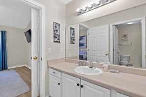 A bathroom with a white sink, beige countertop, and a large mirror with lights. There are two framed pictures on the wall and a blue curtain on the left. - Chapin 2/2 Remodeled in 2025 (Chapin)