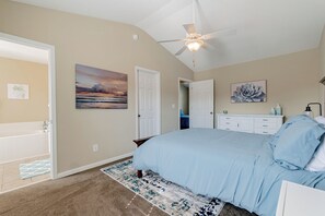 Ein Schlafzimmer mit einem Bett, einem Deckenventilator, einem Bild, einer Kommode und einem durch eine offene Tür einsehbaren Badezimmer.