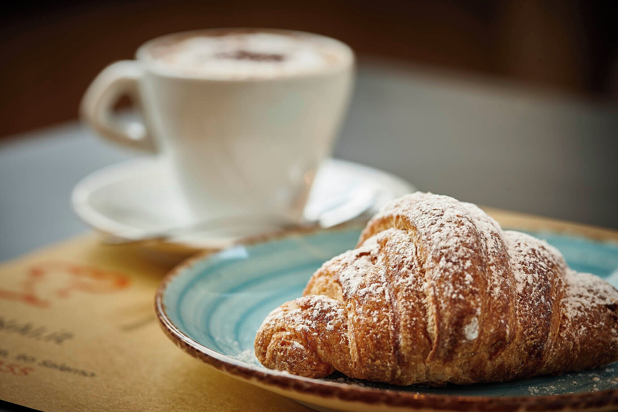 A croissant on a plate with powdered sugar and a cup of coffee on a saucer.