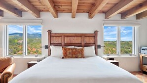 Ein Schlafzimmer mit einem hölzernen Kopfteil, einem großen Bett, zwei Nachttischen, einem Sessel und Blick auf eine gebirgige Landschaft durch große Fenster.