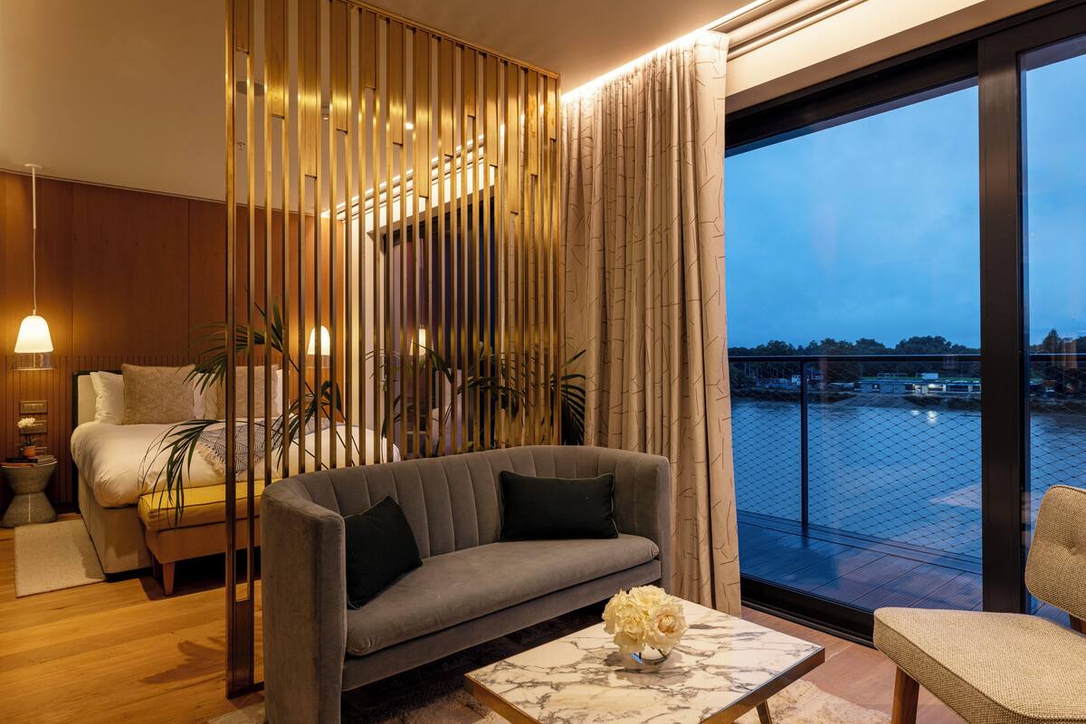 Quarto de hotel moderno com sofá, cama, vista para a água e um painel decorativo.