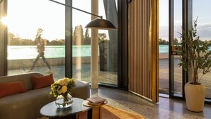A modern room with a sofa, a small table with a vase of flowers, and a large window offering a view of a bridge and greenery. - Fulham Pier Hotel (London)
