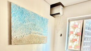 Een kamer met een beige bank, een wandairconditioner en een raam met uitzicht op gebouwen.