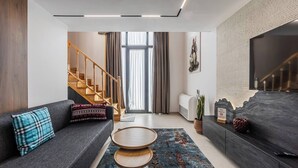 A modern living room with a grey sofa, a small wooden coffee table, a staircase, and a large TV on the wall.