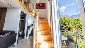 A modern interior with a wooden staircase, a dining area with outdoor seating, and a view of greenery and mountains.
