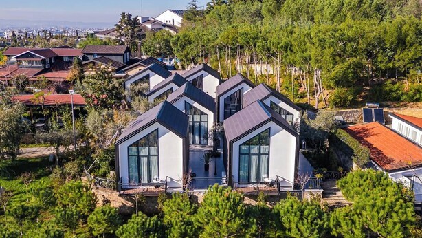 Una casa moderna con un diseño geométrico único, rodeada de abundante vegetación y otras edificaciones residenciales.