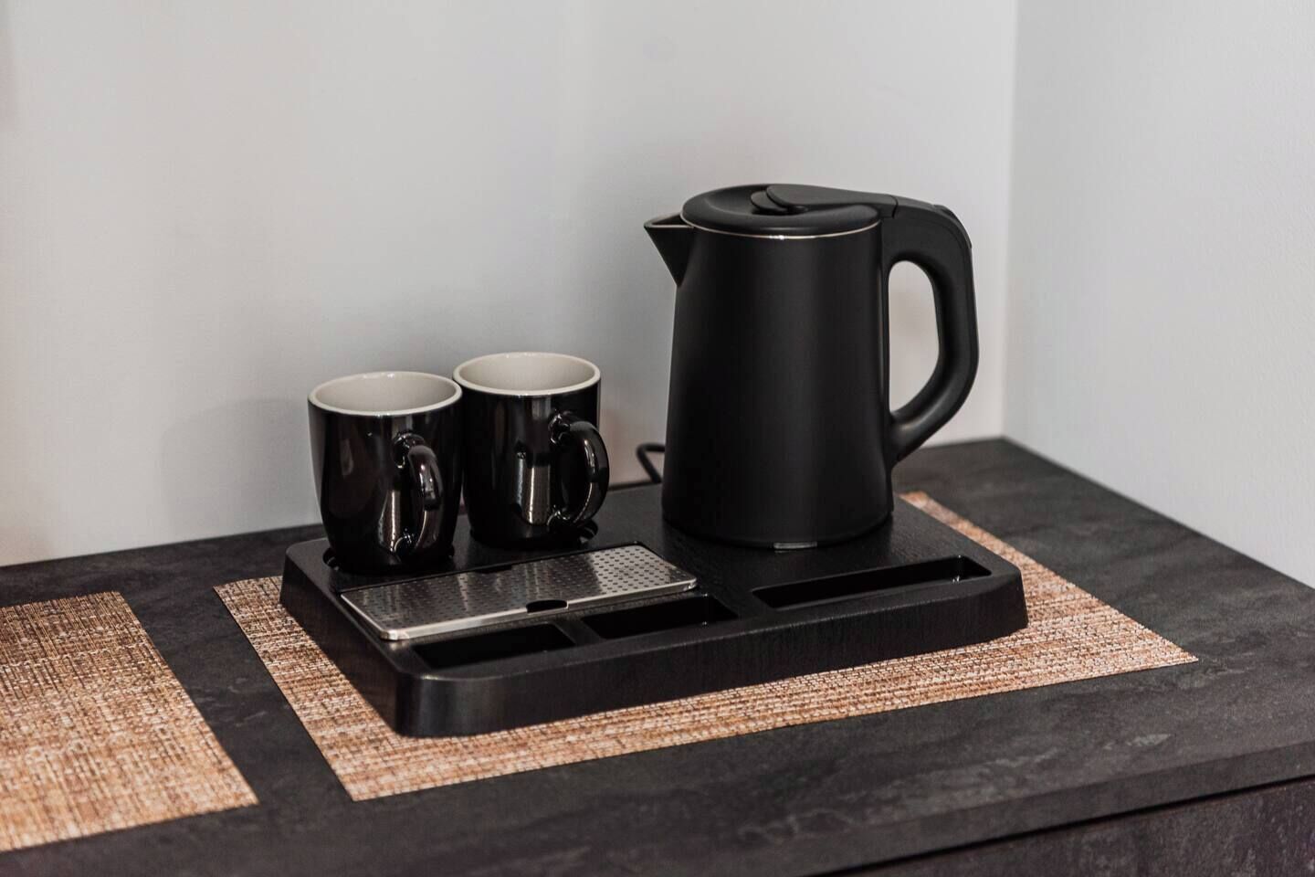 En svart kaffebryggare, två svarta muggar och ett svart bricka på en mörk yta, med en vävd bordsduk under.