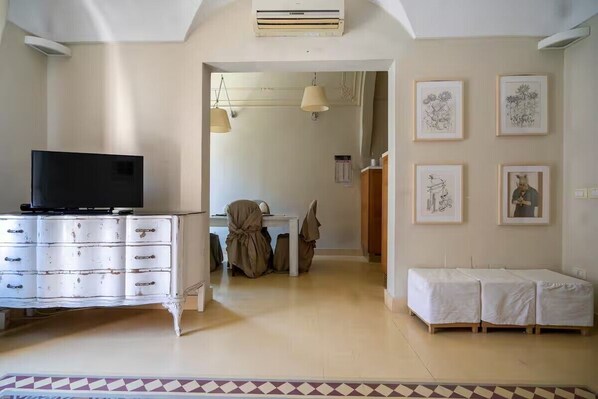 Una habitació amb un cofre blanc, televisió, taula de dinar amb cadires i llit amb roba de llit blanca. Hi ha imatges emmarcades a la paret.