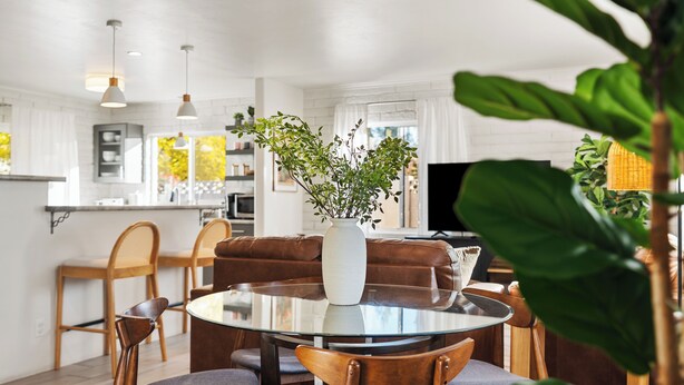 A modern kitchen with white cabinets, a dining area with a glass table and wooden chairs, and a living room with a brown sofa and a large plant. - LOCATION! Uptown Retreat HotTub, Mini Putt, Fire Pit & Views (Sedona)