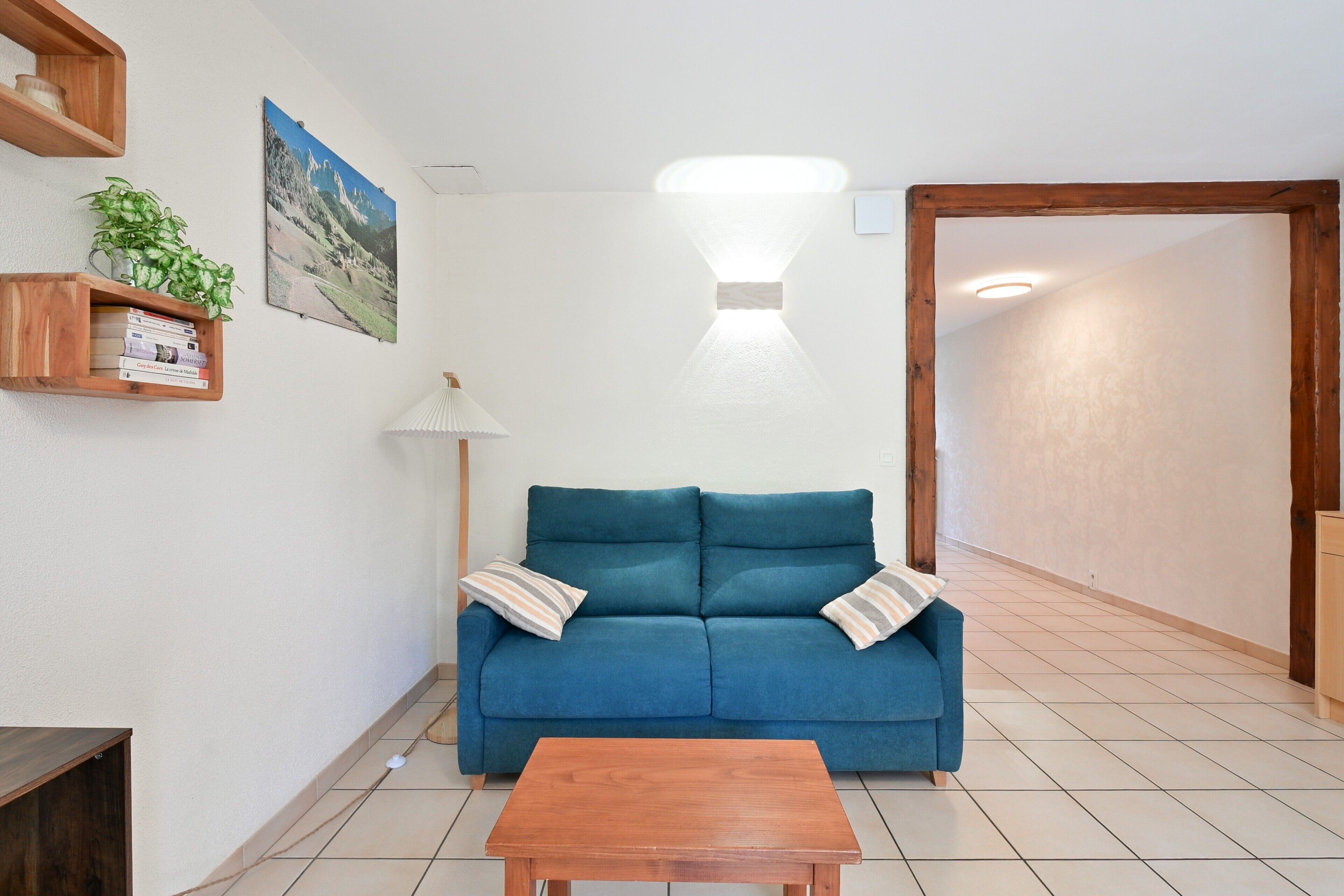 Ein Wohnzimmer mit einem blauen Sofa, einem Holztisch, einer Lampe, einem an der Wand montierten Bücherregal und einem Gemälde.