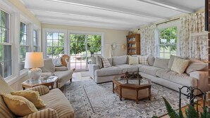 A cozy living room with a large sectional sofa, a coffee table, and a patterned rug. There is a bookshelf and a window with curtains. - Wine Country Farmhouse (Culpeper)
