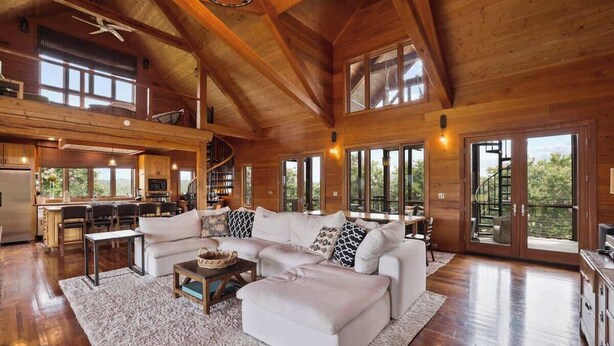 A spacious living area with a large white sectional sofa, a wooden ceiling, and a view of the outdoors through large windows. - The Houses at Lumusa (Culpeper)