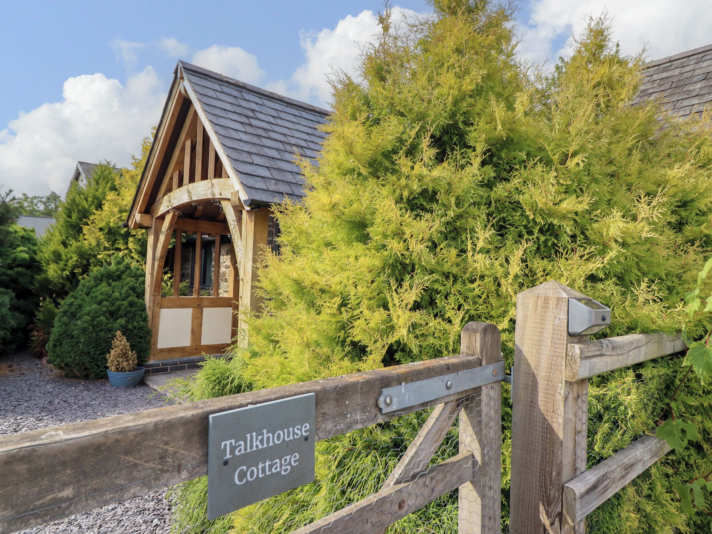 A wooden cottage with a sloped roof, surrounded by dense greenery and a gravel path.