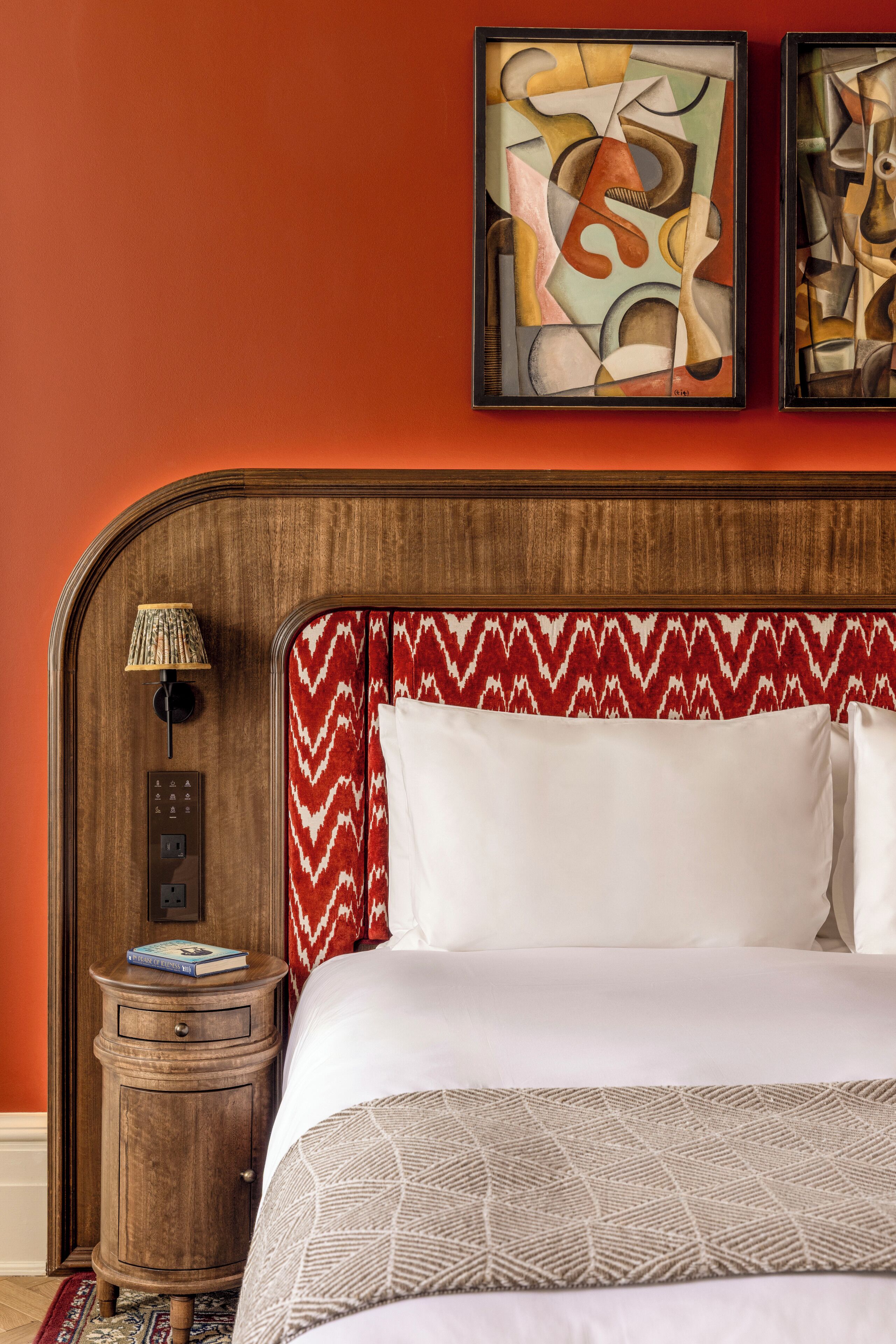 A hotel room with a wooden headboard, a red and white patterned bedspread, a bedside table with a lamp, and abstract art on the wall.