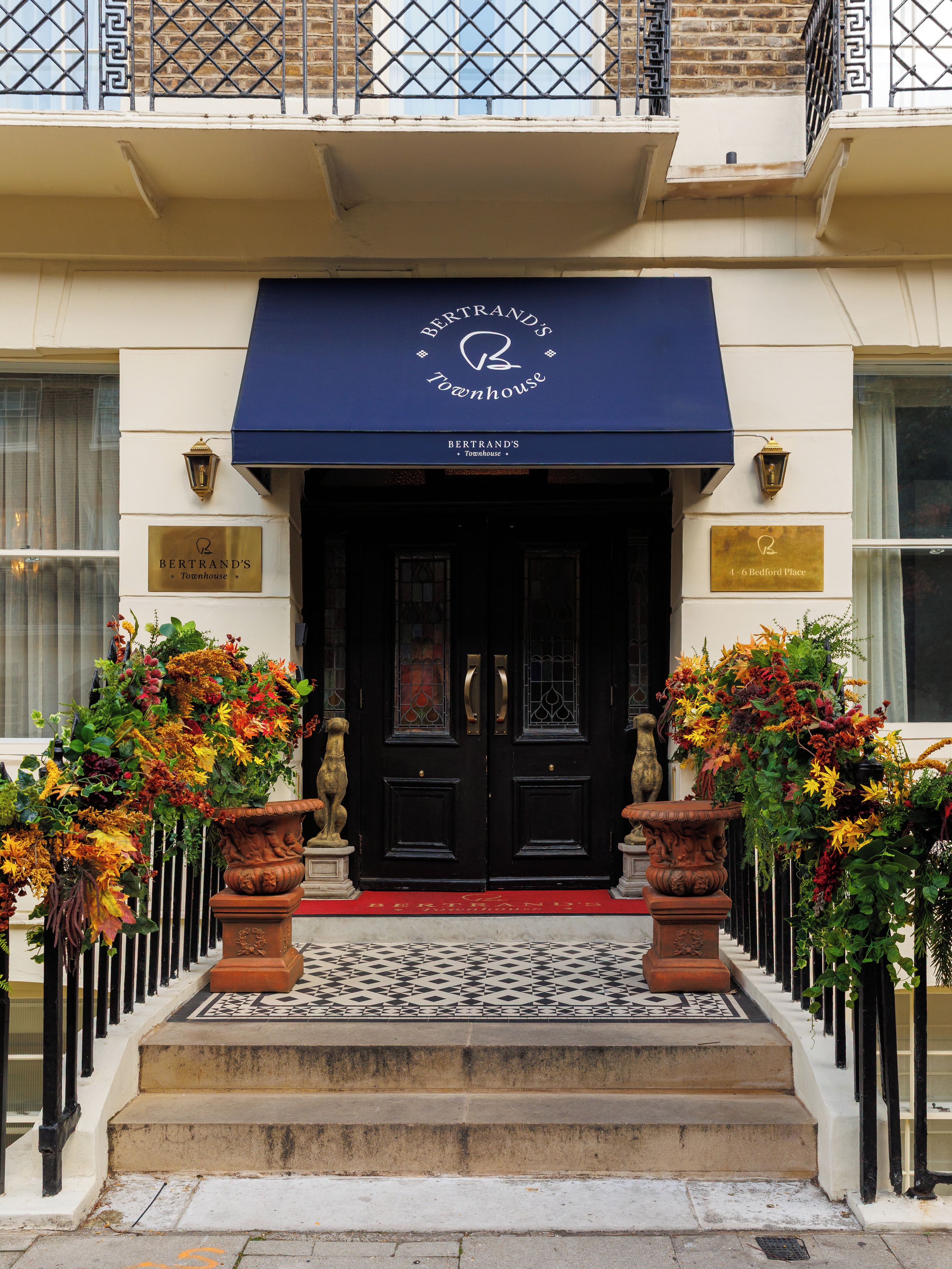 L’entrée d’un immeuble se distingue par un auvent bleu, une porte noire et un tapis rouge menant à l’entrée. De chaque côté de la porte, on peut admirer deux grands arrangements floraux.