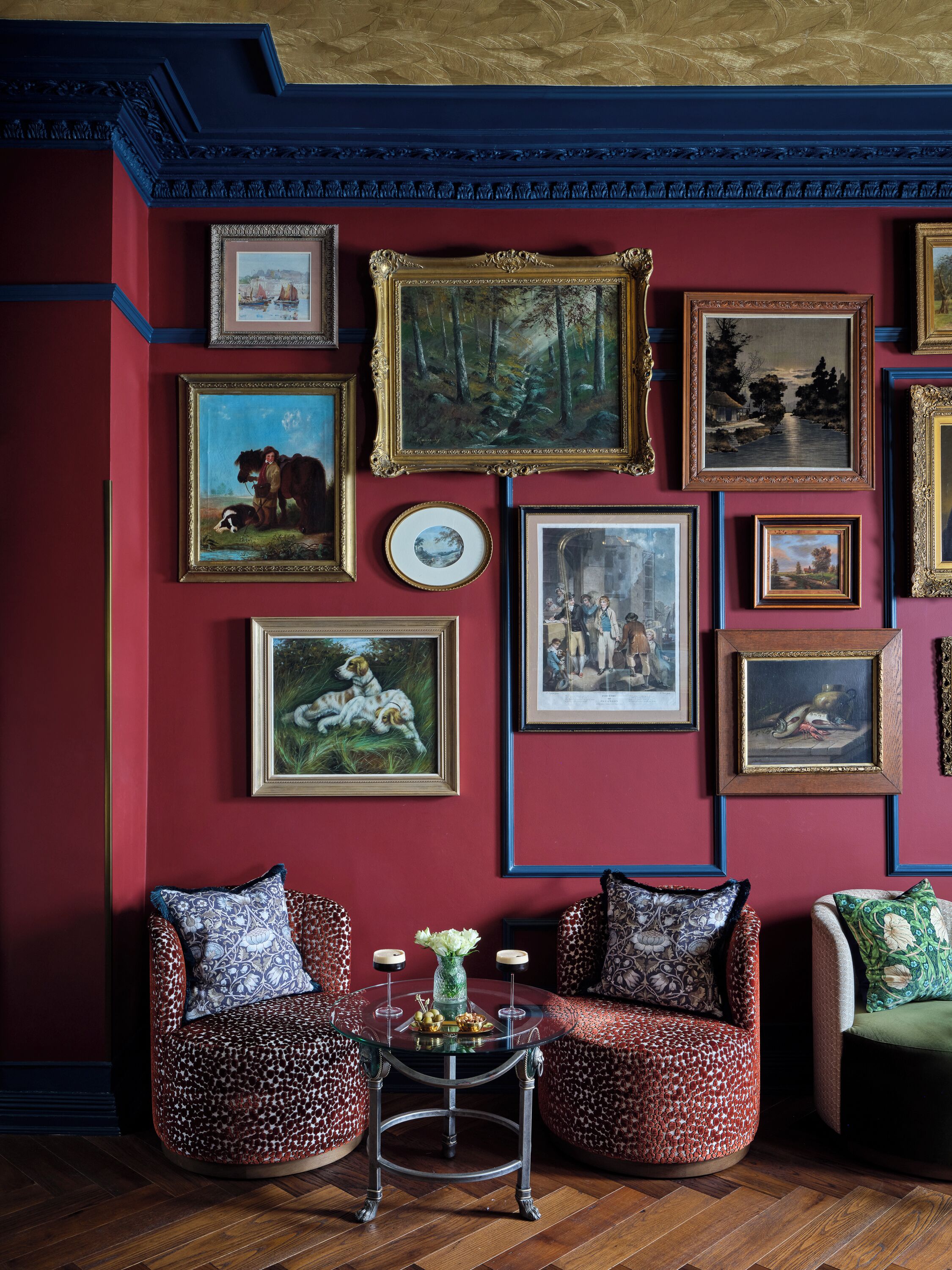 A room with a red wall adorned with various framed artworks, two patterned armchairs, a small round table with candles, and a green upholstered sofa.