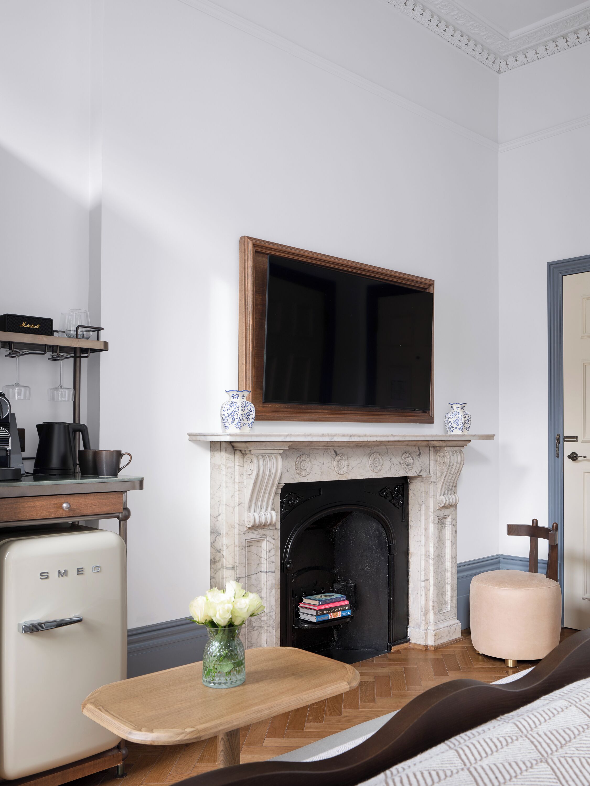 A room with a fireplace, a television mounted above it, a vintage-style refrigerator, a wooden side table with a vase of flowers, and a cushioned chair.