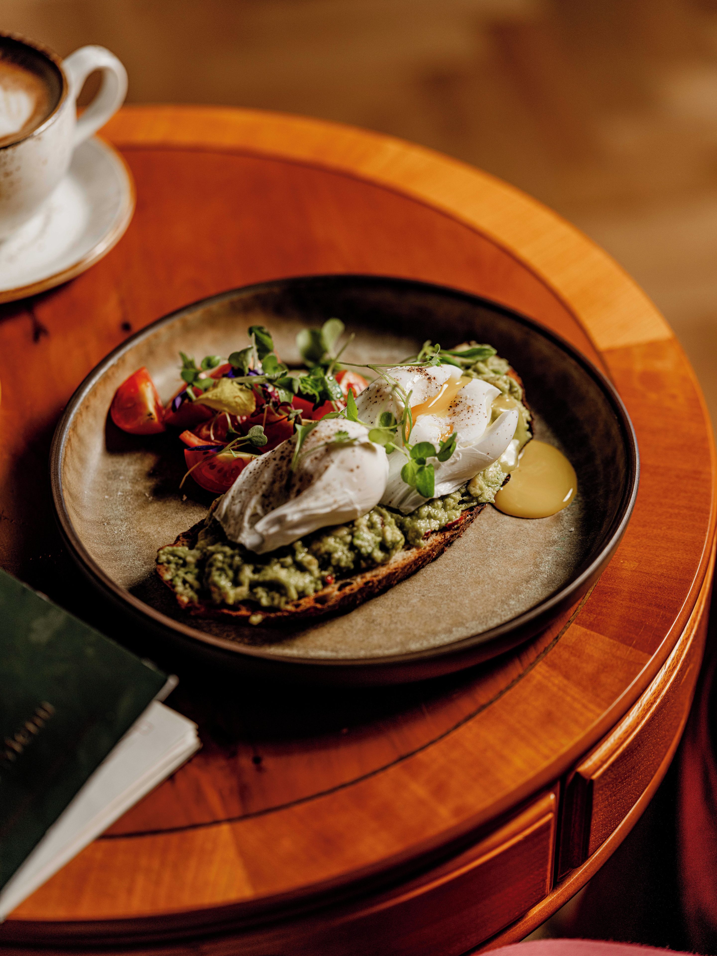 A breakfast plate with poached eggs, avocado spread, and greens on toast, accompanied by a cup of coffee on a wooden table.