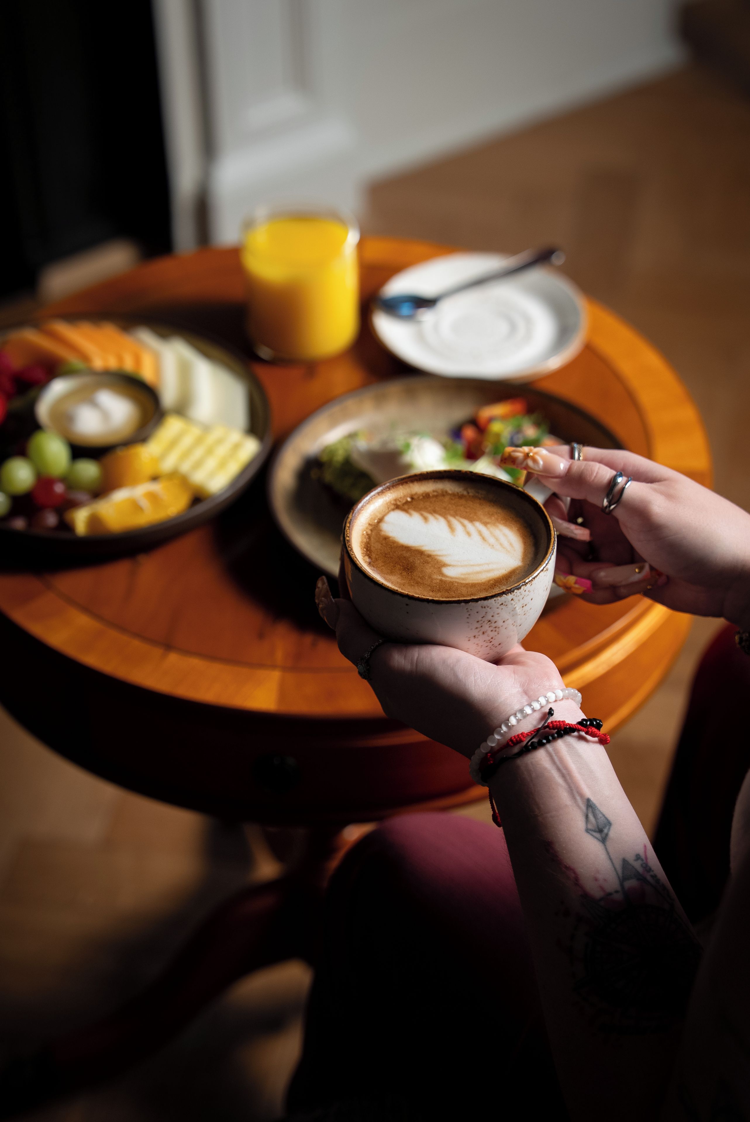 A person holding a cup of coffee with latte art, a plate of food, and a glass of orange juice on a wooden table.