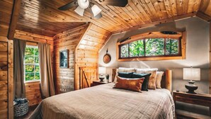 A cozy log cabin bedroom with a large bed, wooden walls, a ceiling fan, and a window with a view of trees. - NEW! The Cowboy-5BR Cabin w/HT, Game Room & Pool Access (Sevierville)
