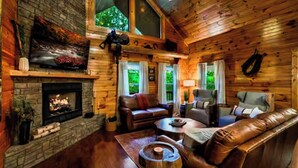 A log cabin interior with a stone fireplace, leather furniture, and a large window overlooking greenery. - NEW! The Cowboy-5BR Cabin w/HT, Game Room & Pool Access (Sevierville)