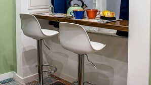 A small kitchenette with two white bar stools, a window with blue curtains, and a tiled floor. - Inner Naples (Naples)