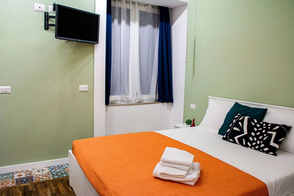 A hotel room with a bed, a flat-screen TV mounted on the wall, a window with sheer curtains, and a patterned floor.