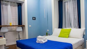 A neatly made bed with a blue bedspread and a green pillow, a small table with a yellow pitcher, and a chair with a white cushion. - Inner Naples (Naples)