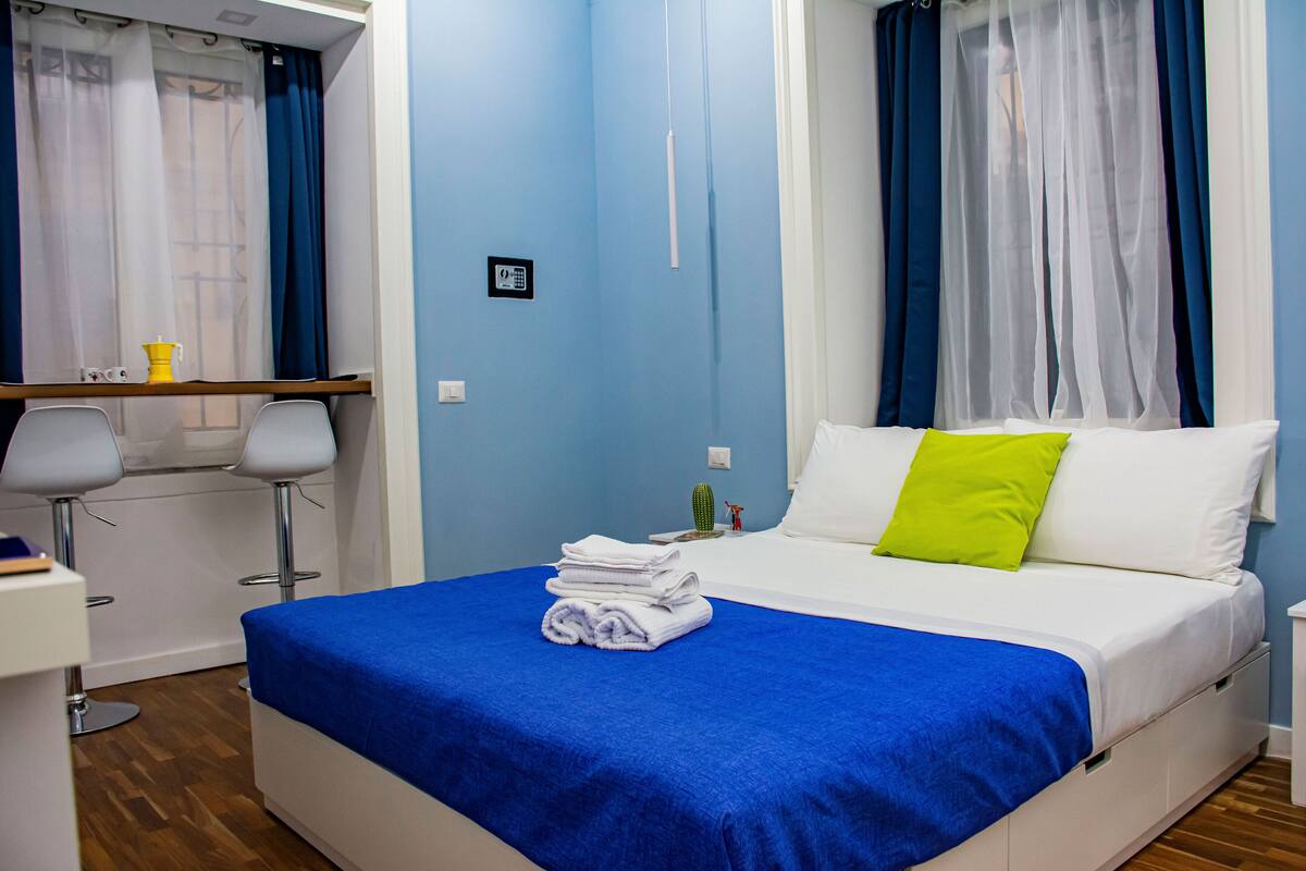 A neatly made bed with a blue bedspread and a green pillow, a small table with a yellow pitcher, and a chair with a white cushion.
