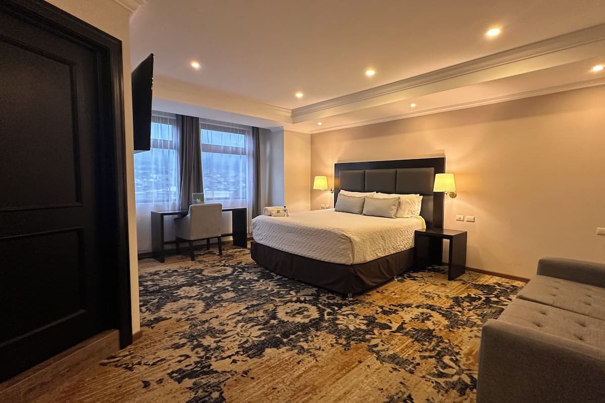 Une chambre d’hôtel dotée d’un grand lit, de deux tables de chevet avec des lampes, d’un bureau avec une chaise et offrant une vue sur la ville grâce à de grandes fenêtres.
