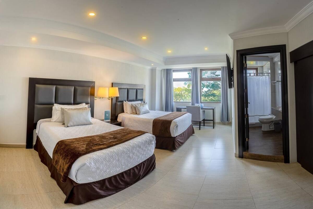 Une chambre d’hôtel avec deux lits, un bureau et une salle de bain visible à travers une porte ouverte.