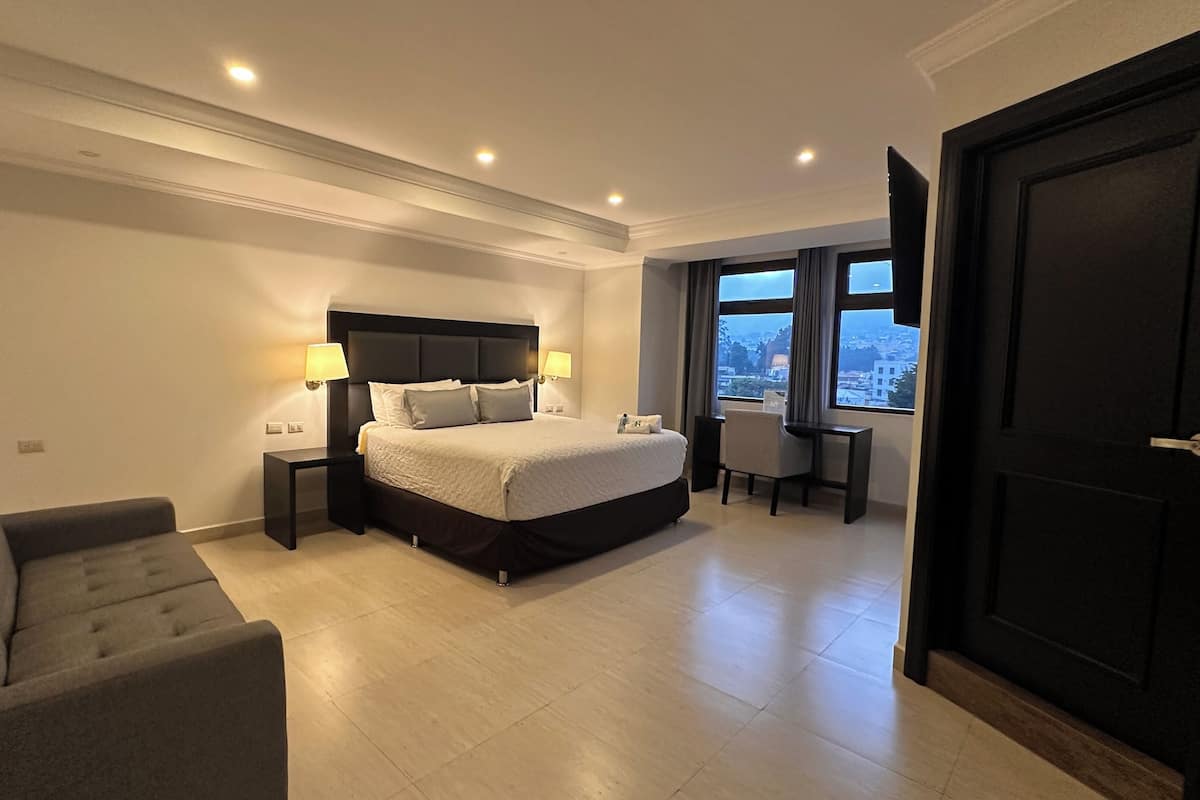 Une chambre d’hôtel dotée d’un grand lit, de tables de chevet avec des lampes, d’un canapé, d’un bureau et offrant une vue sur la ville grâce à de grandes fenêtres.