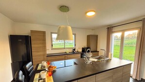 A modern kitchen with a large island, wooden cabinets, and a view of a green landscape through the window. - La Maison du Pinier – Nature (Royères)