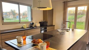 A modern kitchen with a large island, a coffee maker, a toaster, and a view of a garden through the windows. - La Maison du Pinier – Nature (Royères)
