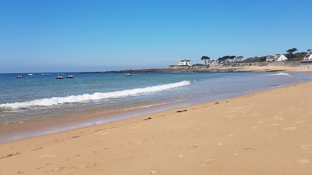 A sandy beach with waves, a clear sky, and buildings in the background.