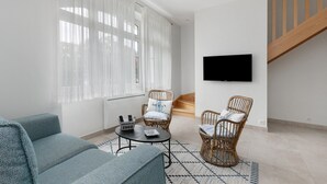 A modern living room with a sofa, two wicker chairs, a coffee table, and a flat-screen TV mounted on the wall.