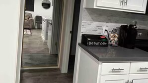Uma cozinha moderna com armários brancos, um espaço para café e vista para uma sala de estar com sofá e lareira.