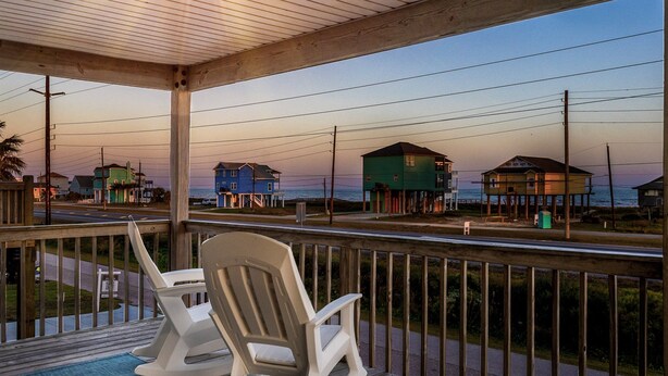 Um deck de madeira com duas cadeiras de balanço e vista para as casas na praia ao entardecer.