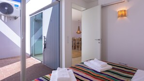 A hotel room with a sliding glass door leading to an outdoor area, a bed with a striped blanket, a bedside table, and a wall-mounted air conditioner. - CASA DO REGADIO - CASA 3, A country and sea retreat on the Alentejo coast, (Beja)
