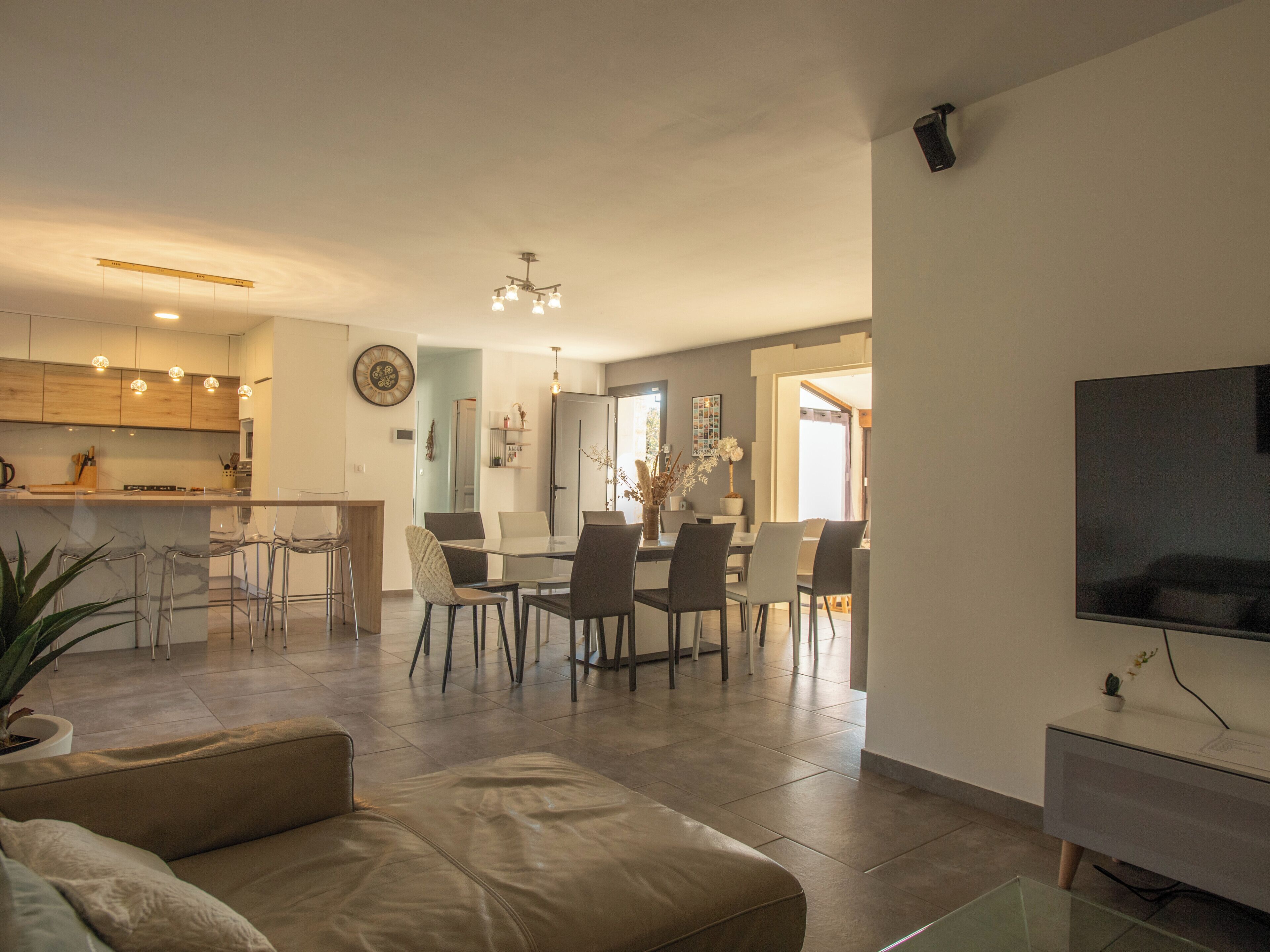 En moderne stue med sofa, spiseområde med stole, et køkken med træskabe og et fjernsyn på væggen.