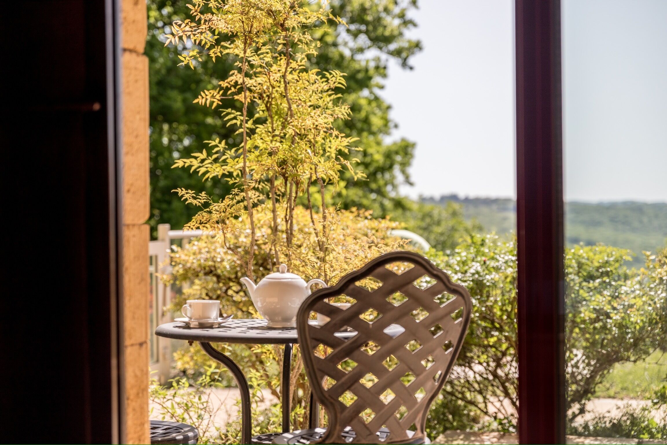 A table with a teapot and cup, a chair, and a view of greenery outside.