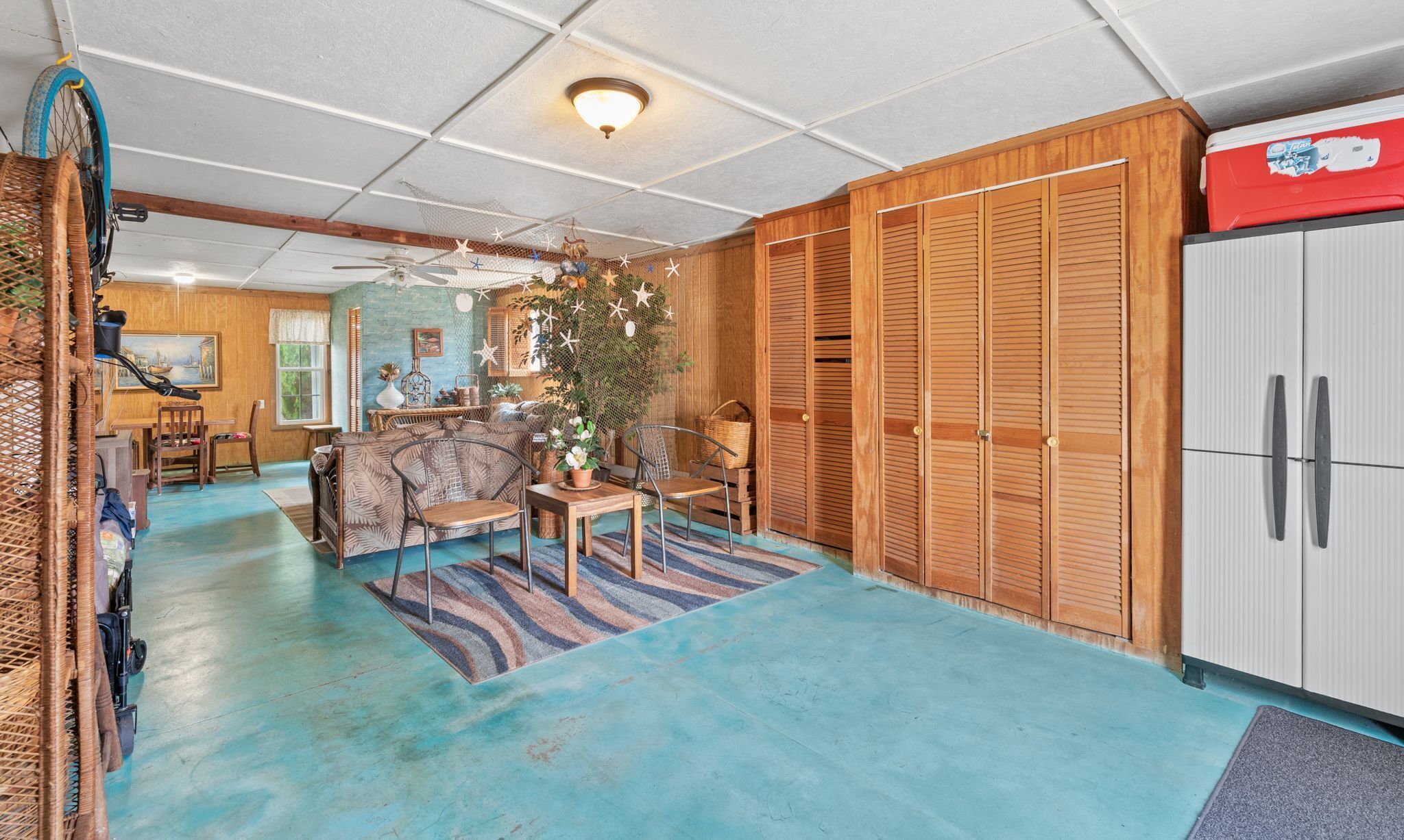 A room with a blue floor, a wooden wardrobe, a dining area with a table and chairs, and a ceiling fan.