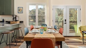 A dining area with a wooden table set for four, surrounded by chairs, with a view of trees through large windows. - Family Friendly Modern Cabin w/ Hot Tub, Fire Pit & EV Charger - Lost River WV (Lost City)