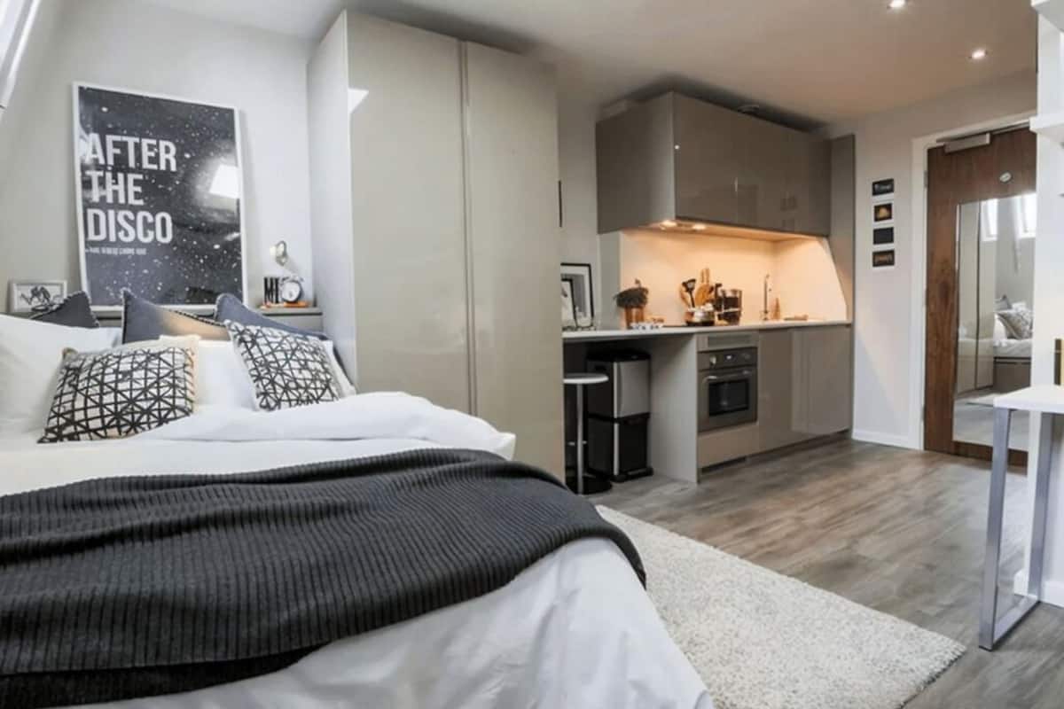 A modern bedroom with a large bed, a small kitchenette, and a bathroom visible through an open door.