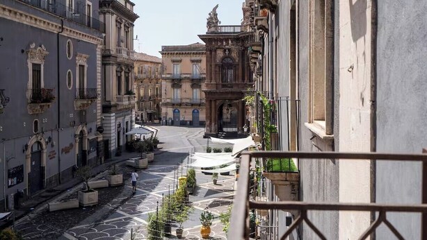 Una calle estrecha flanqueada por edificios, un espacio abierto central con plantas en macetas y una barandilla de balcón en primer plano.
