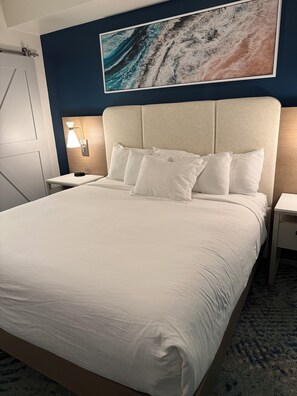 A neatly made bed with white linens, a headboard with a light wood finish, and a wall-mounted artwork depicting a wave. - Rare, Luxury, 3 Bedroom/2 Bathroom Oceanfront Condo in Myrtle Beach, SC (Myrtle Beach)