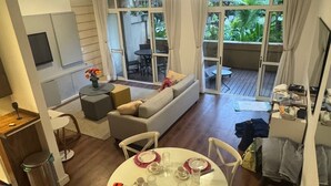 Un coin repas avec une table ronde dressée pour quatre personnes, entourée de chaises blanches. On aperçoit, au fond, un canapé et une petite table avec des chaises, ainsi qu’une vue sur un balcon agrémenté de plantes.