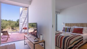 Une chambre d’hôtel moderne dotée d’une grande porte-fenêtre coulissante donnant sur un patio extérieur, d’un lit avec une literie à rayures et d’un téléviseur posé sur une table en bois.